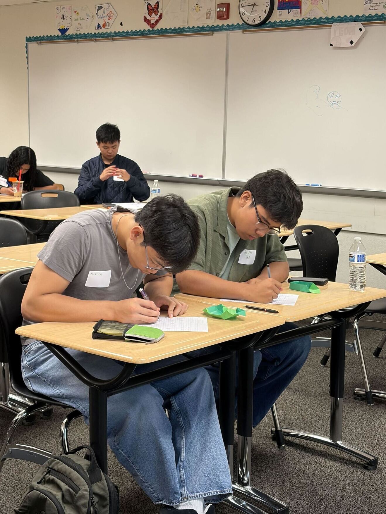 Two students writing on notebook paper. They both have a green origami box in front of them.