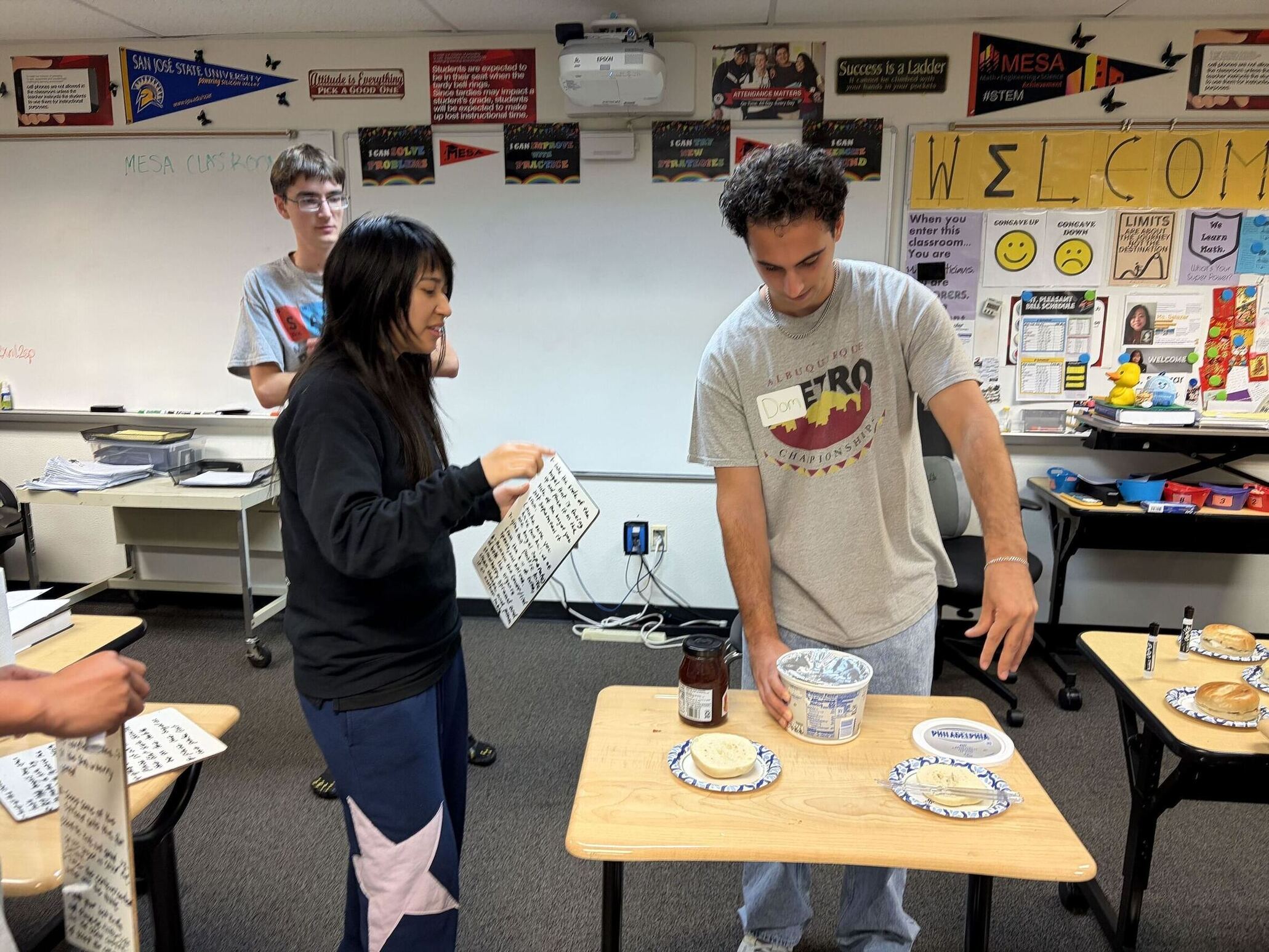 Three people. In front of one of them is a split bagel and container of cream cheese.